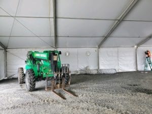 Inside a large, white industrial tent with a green telescopic handler on gravel ground, suggesting an active construction site.