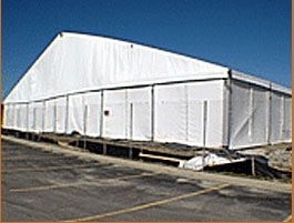 Large white temporary structure with a pitched roof on asphalt, indicative of a commercial tent or temporary warehouse.